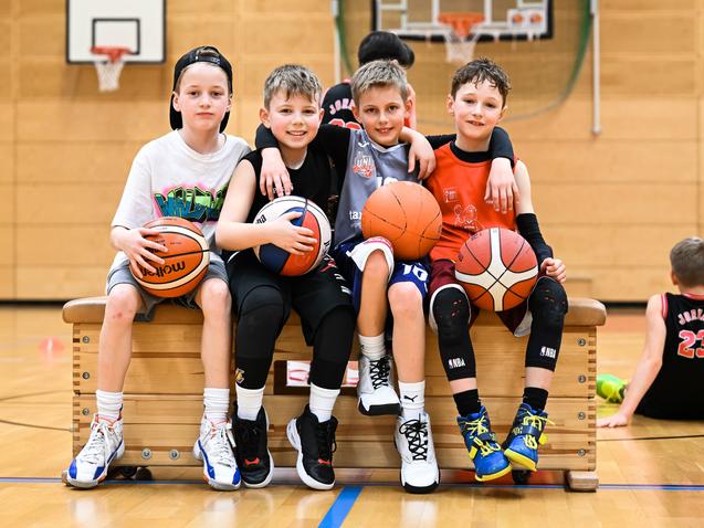 Vier Kinder sitzen auf einer Bank in der Turnhalle, halten Basketballs und lachen. Im Hintergrund sind weitere Kinder aktiv.