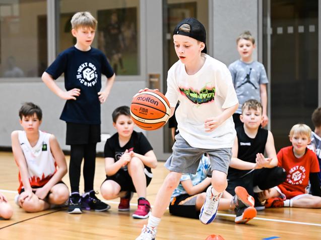 Ein Junge dribbelt einen Basketball auf einem Sportboden, während andere Kinder im Hintergrund zuschauen.