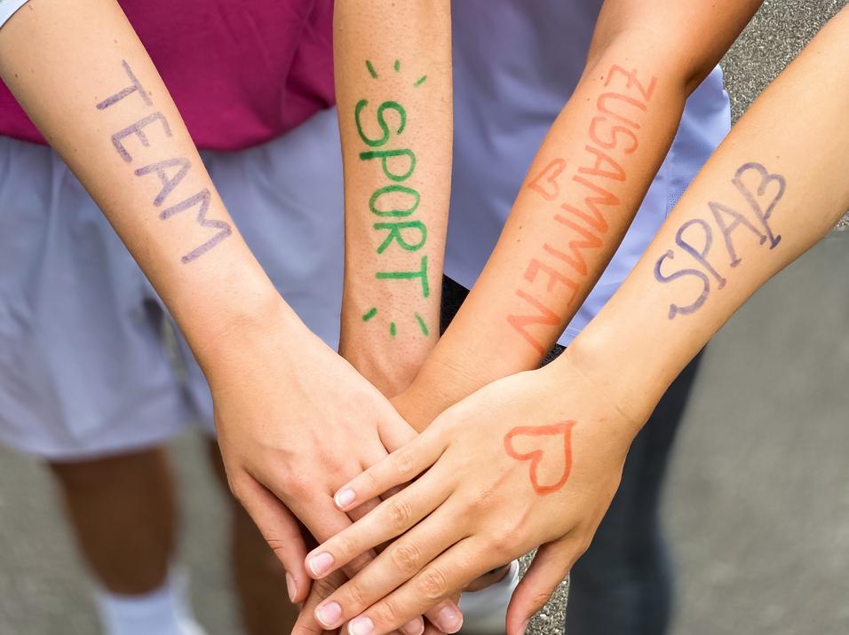 Vier Hände mit bunten Schriftzügen "TEAM", "SPORT", "ZUSAMMEN" und "SPASS" liegen übereinander auf einem grauen Untergrund.