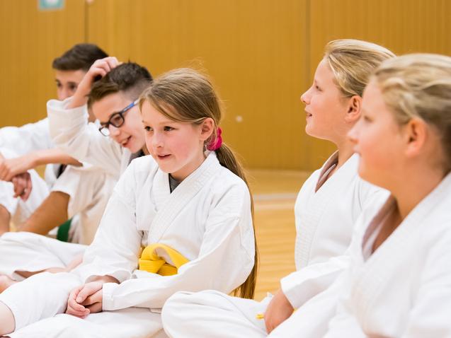 Fünf Kinder in weißen Karateanzügen sitzen in einer Trainingshalle und unterhalten sich.