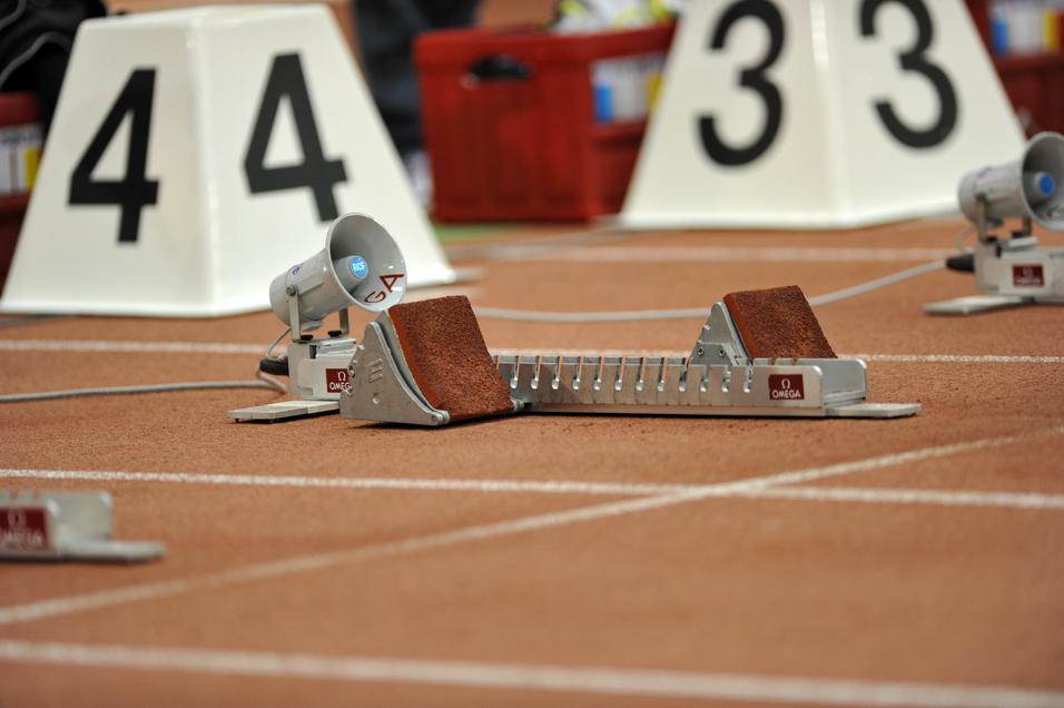 Startblöcke und Zeitmessgeräte auf einer Leichtathletikbahn mit brauner Oberfläche und großen Wettkampfnummern im Hintergrund.